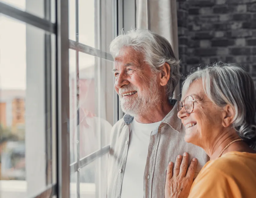 Pareja de ancianos mirando por la ventana de su casa mientras sonríen